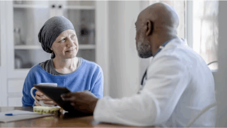 A physician in a white coat speaks with an older woman in a blue shirt with a scarf over her head, who appears to have undergone cancer treatment.